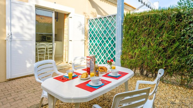 Un espace de restauration en plein air avec une table dressée pour deux personnes, entourée de chaises en plastique blanches, d’un parasol blanc et d’une clôture en treillis vert.