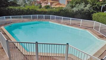 Une piscine rectangulaire entourée d’une clôture blanche, avec de la verdure tout autour et des bâtiments en arrière-plan.