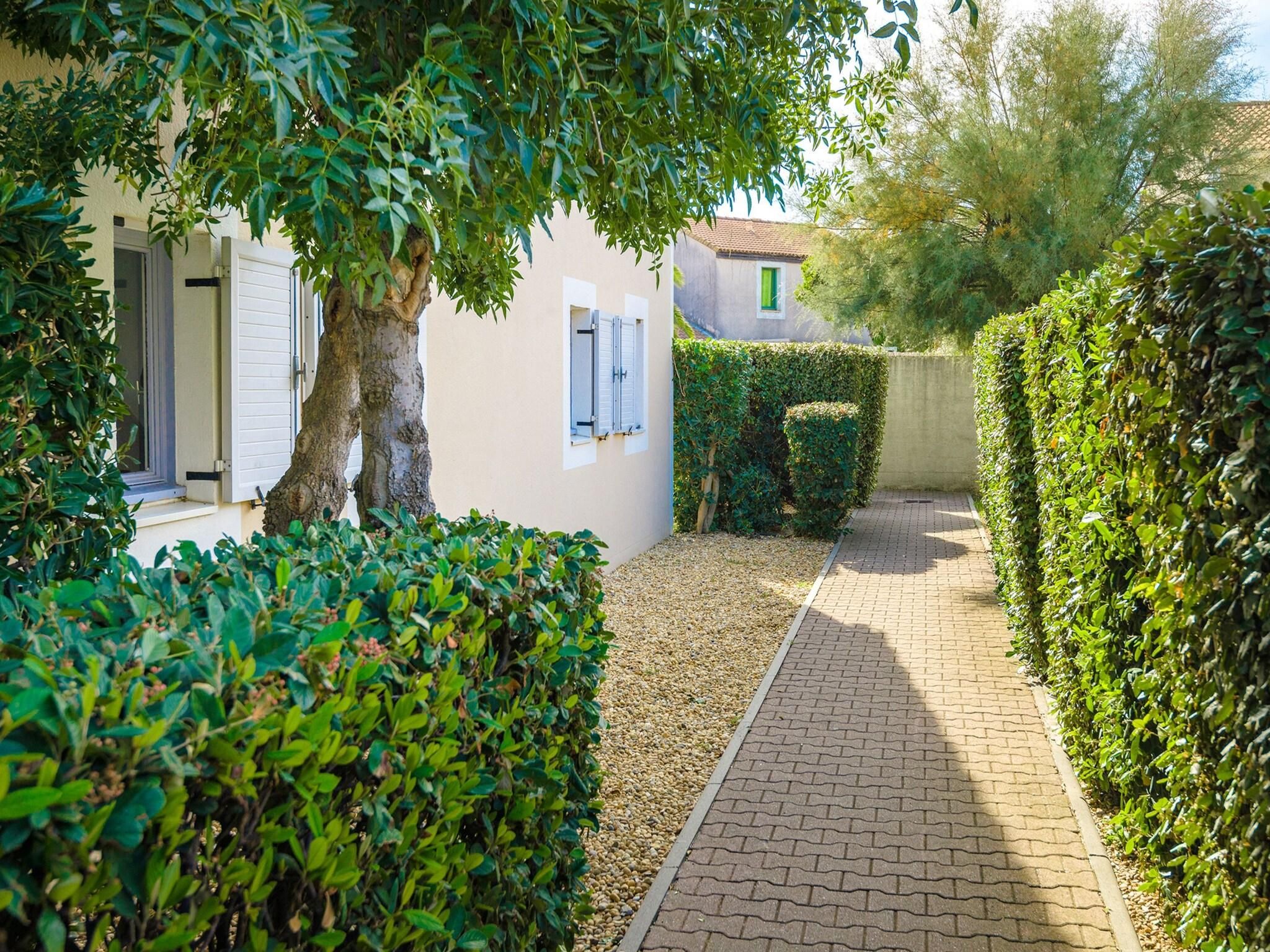 Un étroit sentier pavé, bordé de haies soigneusement taillées, et un bâtiment à la façade blanche et aux fenêtres fermées par des volets.