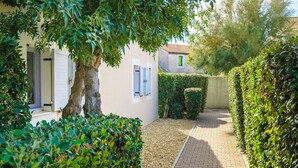 A narrow paved pathway flanked by neatly trimmed hedges and a building with a white facade and shuttered windows. - Residence Les Soleillades Et Scarlett (Palavas-les-Flots)