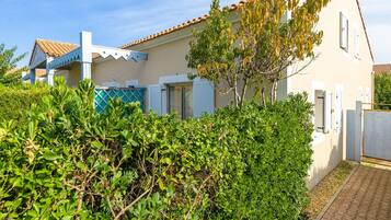Une maison à deux étages, avec un toit en tuiles, un balcon et un jardin où poussent différentes espèces de plantes.
