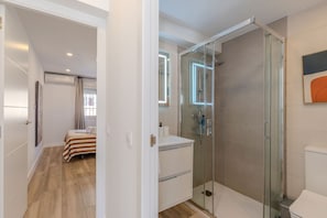 Uma casa de banho moderna com cabine de duche em vidro, lavatório branco e uma cama no quarto vizinho.