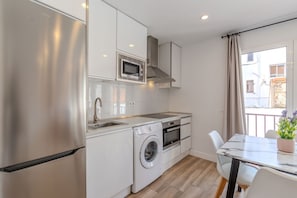 Cozinha compacta com frigorífico de aço inoxidável, micro-ondas integrado, máquina de lavar roupa e área de refeições com mesa e cadeiras.