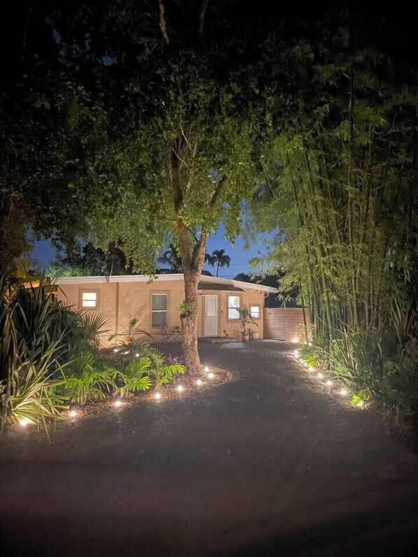 A house with a paved driveway, a tree in the center, and a small garden with plants and lights.