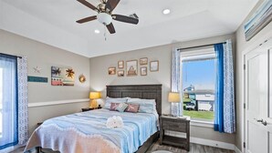 Ein Schlafzimmer mit Bett, Nachttischen, Deckenventilator, Fenster mit Vorhängen und Wandbildern.