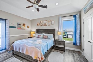 Ein Schlafzimmer mit Bett, Nachttischen, Deckenventilator, Fenster mit Vorhängen und Wandbildern.