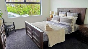 Un dormitorio con un ventanal grande, una cama con ropa de cama estampada, una mesita de noche con lámpara y una silla.