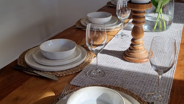 Una mesa de comedor decorada con platos, cuencos y copas de vino blancos, una vela y un florero con flores.