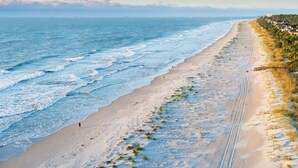 Uma praia com vista clara do oceano, um caminho de areia e uma fileira de edifícios à beira-mar.