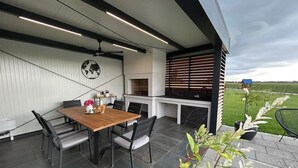 A modern outdoor dining area with a wooden table and chairs, a built-in grill, and a view of the countryside. - Riverside retreat with pool, summer kitchen, playground, and peaceful Drava views. (Pitomača)