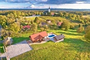 Un paysage rural avec une grande maison dotée d’une piscine, entourée d’une végétation luxuriante et d’une église visible au loin.