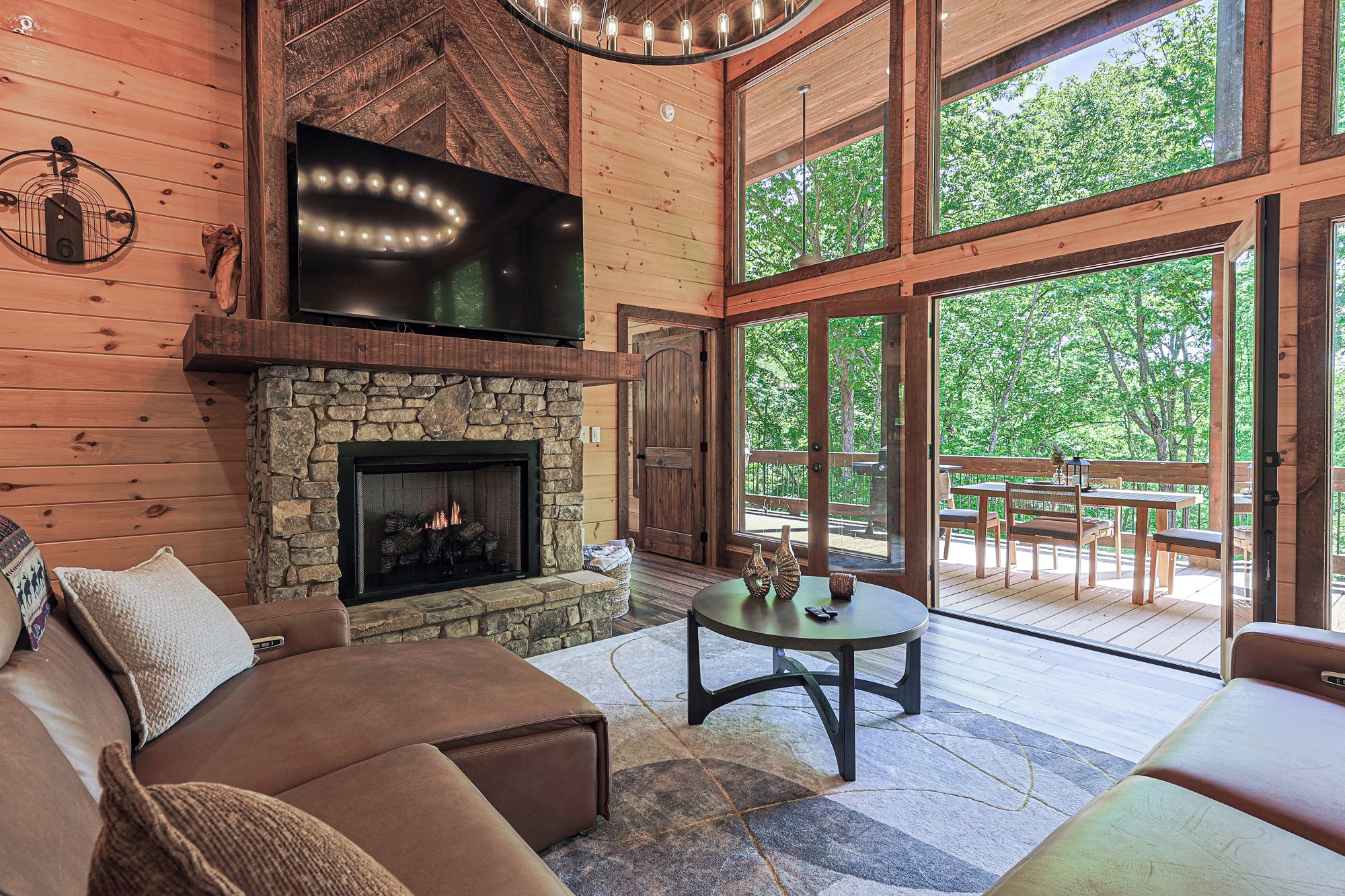 A cozy interior with a stone fireplace, wooden walls, and large windows offering a view of the outdoors.