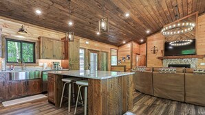 A rustic cabin interior with a wooden ceiling, a stone fireplace, a large TV, and a bar area. - Rugged Trail Oasis: Modern 3BR Cabin w/ 3 Fireplaces & Mountain Views (Morganton)