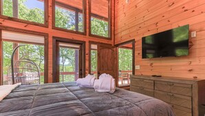 A wooden cabin bedroom with a large bed, a TV mounted on the wall, and a view of the outdoors. - Rugged Trail Oasis: Modern 3BR Cabin w/ 3 Fireplaces & Mountain Views (Morganton)