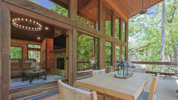 A wooden deck with a table and chairs, a stone fireplace, and large windows overlooking a forest. - Rugged Trail Oasis: Modern 3BR Cabin w/ 3 Fireplaces & Mountain Views (Morganton)