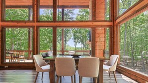 A dining area with a table and chairs, large windows, and a view of trees. - Rugged Trail Oasis: Modern 3BR Cabin w/ 3 Fireplaces & Mountain Views (Morganton)