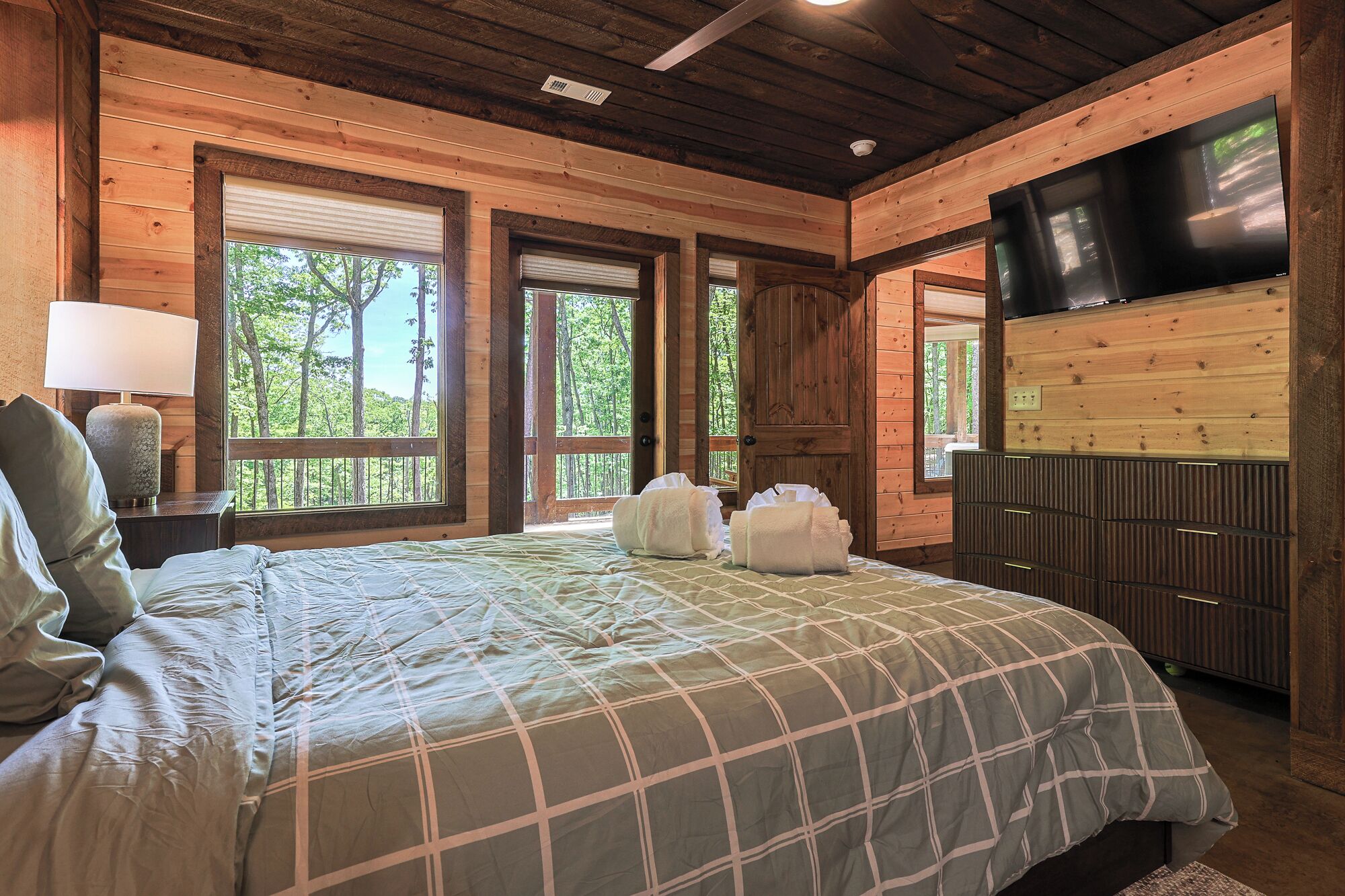 A wooden cabin bedroom with a bed, a TV, and windows with views of trees.