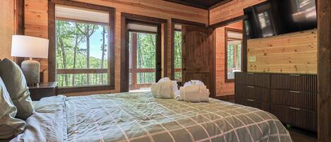 A wooden cabin bedroom with a bed, a TV, and windows with views of trees.