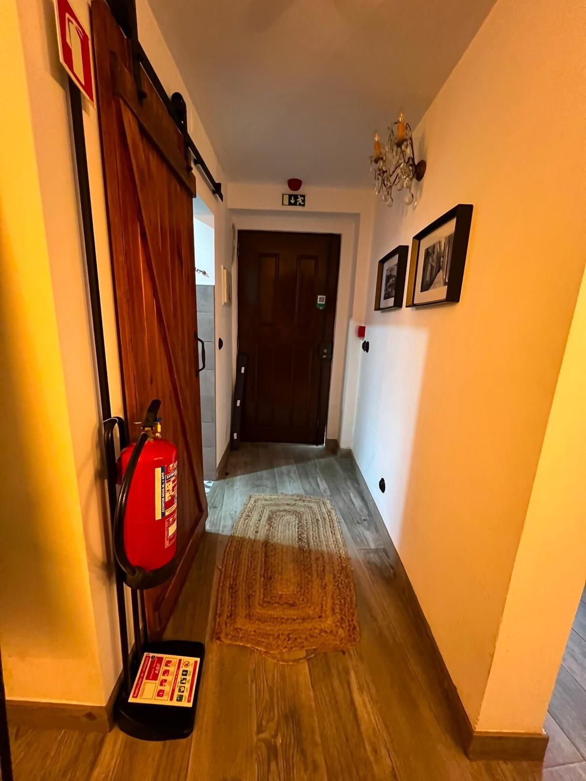 A hallway with a wooden floor, a fire extinguisher mounted on the wall, a door at the end, and a chandelier on the ceiling.