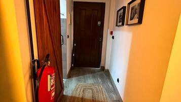 A hallway with a wooden floor, a fire extinguisher mounted on the wall, a door at the end, and a chandelier on the ceiling.