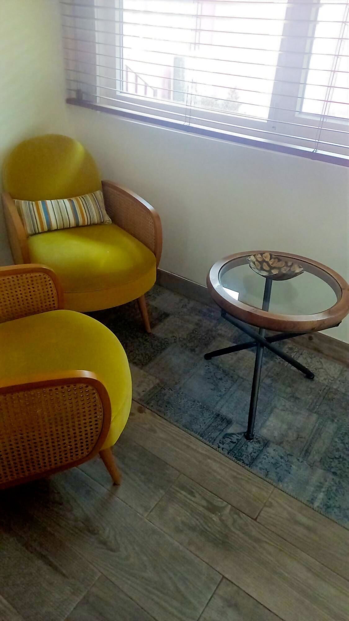 A room with a yellow armchair, a round table with a glass top, and a window with blinds.