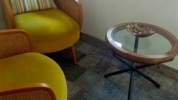 A room with a yellow armchair, a round table with a glass top, and a window with blinds.