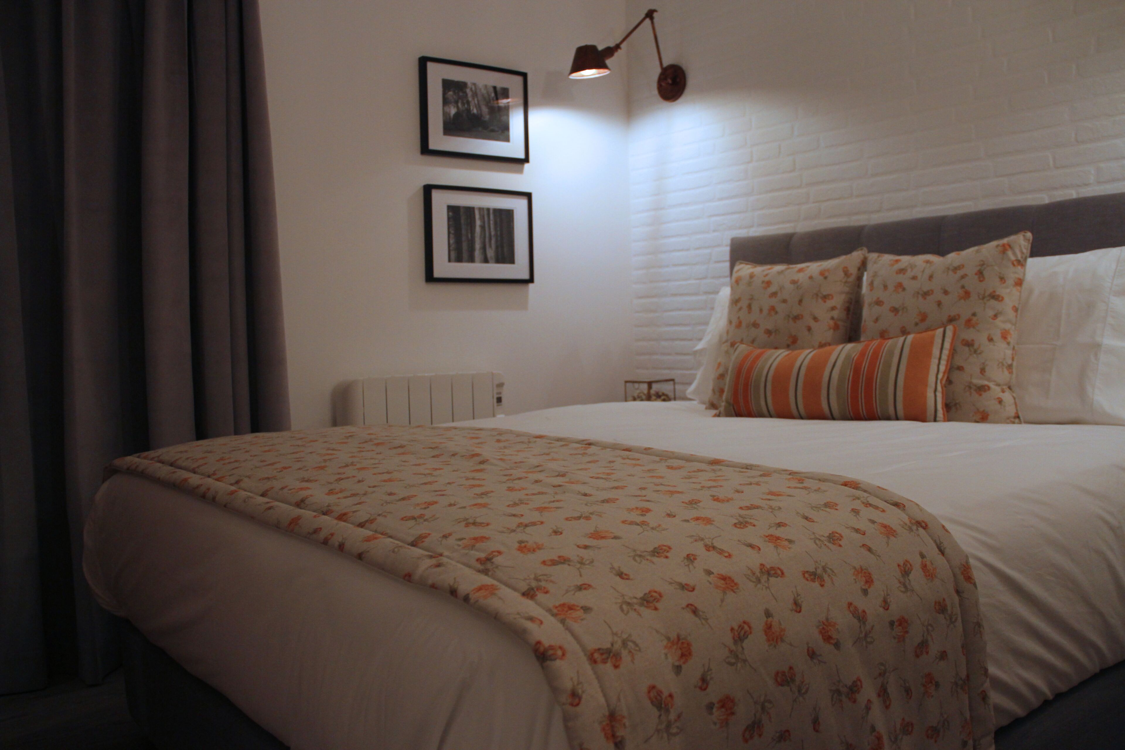 Een net opgemaakt bed met een bloemenprintdeken, een hoofdbord met decoratieve kussens en twee ingelijste foto's aan de muur.