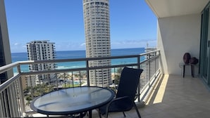 Uma varanda com mesa e cadeiras, com vista para uma praia e oceano, com edifícios altos ao fundo.