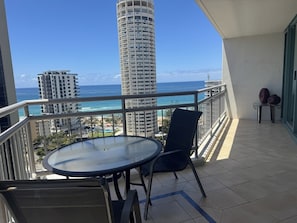 Un balcón con mesa y sillas, con vista a una playa y al océano, y altos edificios al fondo.
