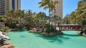 Área de piscina de resort com água turquesa cristalina, rodeada por palmeiras e rochas, com edifícios altos ao fundo.