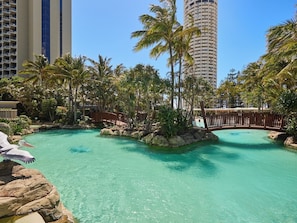 Área de piscina en un resort, con agua turquesa cristalina, rodeada de palmeras y rocas.
