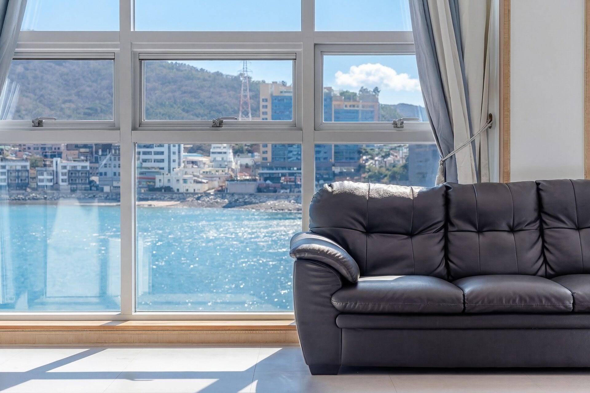 Quarto com uma grande janela, um sofá de couro preto e vista para uma cidade costeira.