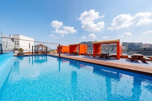 Piscina no terraço com água azul cristalina, áreas de estar em tons de laranja e vista para a paisagem urbana.