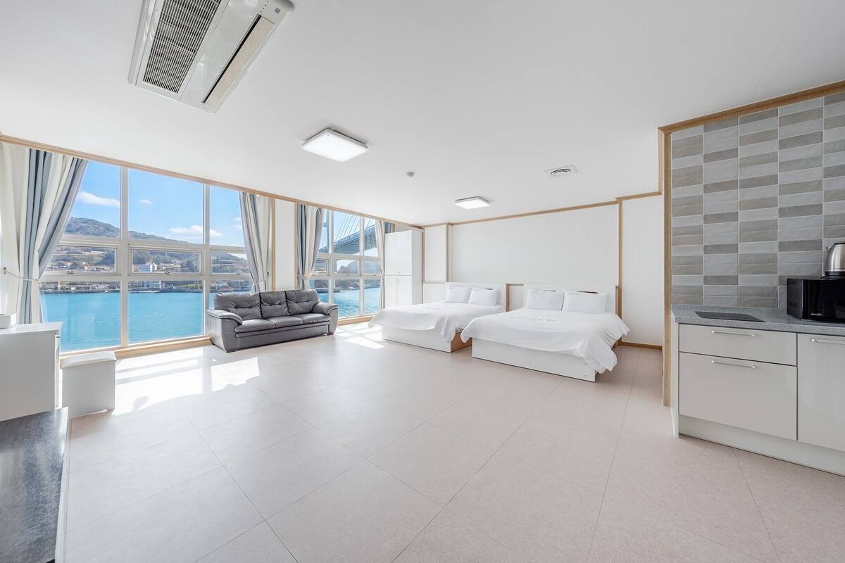 Ein modernes Hotelzimmer mit großem Fenster, zwei Betten, einer Sitzecke und einer Küchenzeile, mit Blick auf eine Brücke und Wasser.