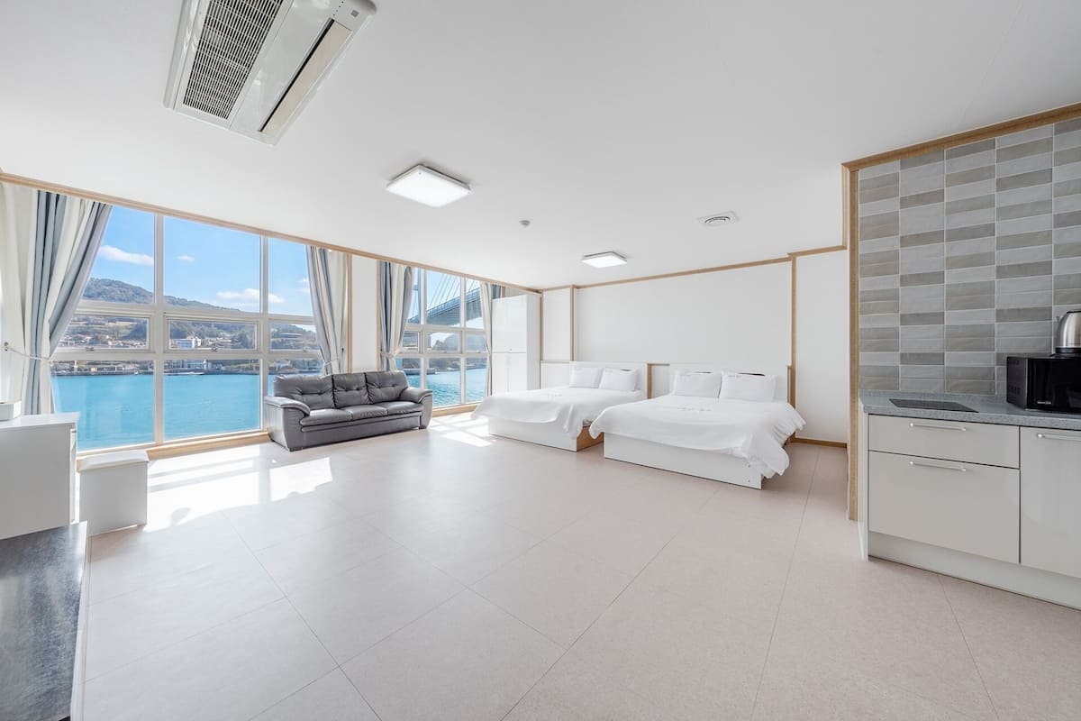 Habitación de hotel moderna con un ventanal grande, dos camas, un sofá, una zona de cocina y vistas a un puente y al agua.