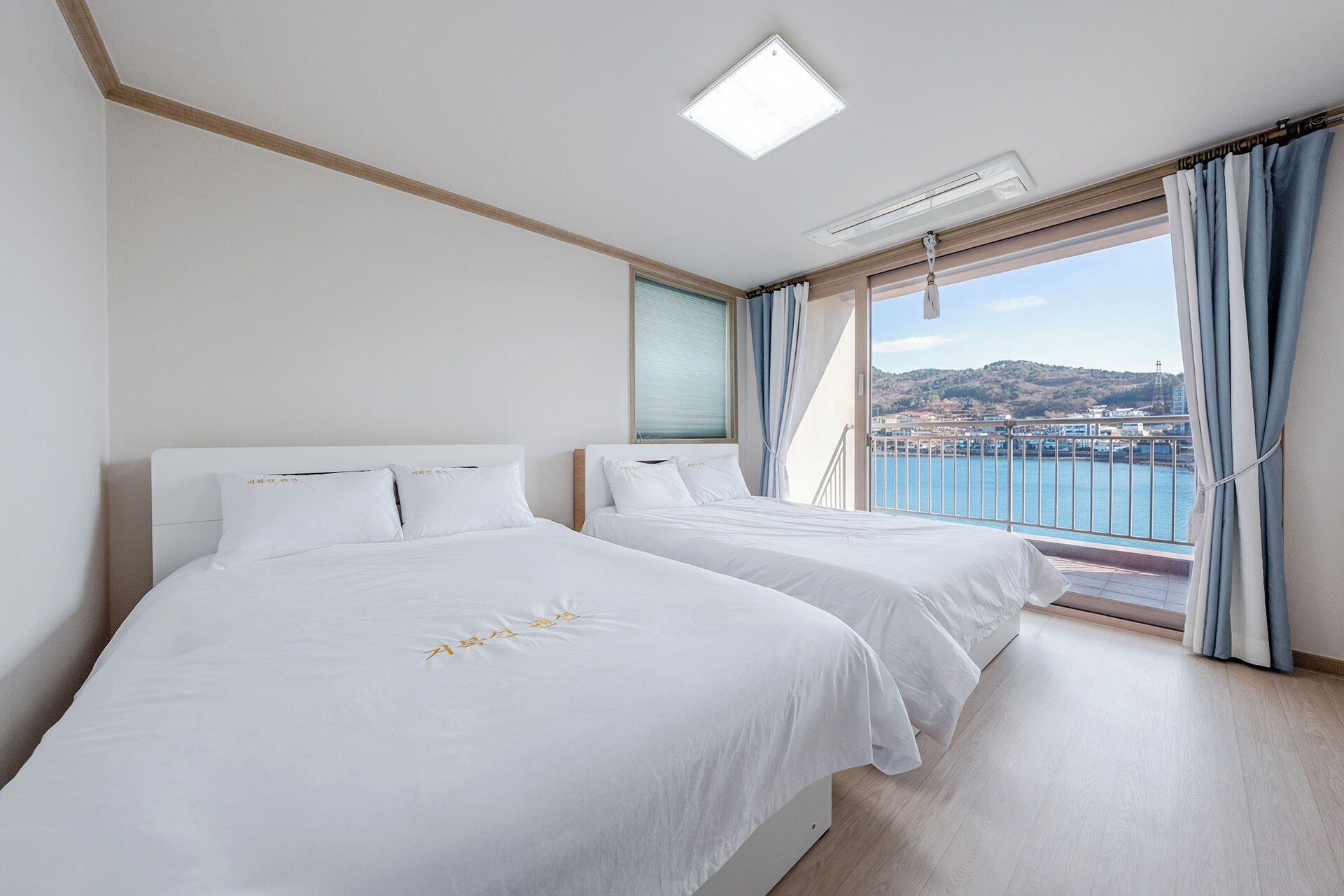 A hotel room with two beds, a large window with a view of a body of water and a cityscape, and a ceiling light fixture.