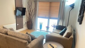 Una sala de estar con dos sofás, una mesa de centro, un televisor y un ventanal con persianas.