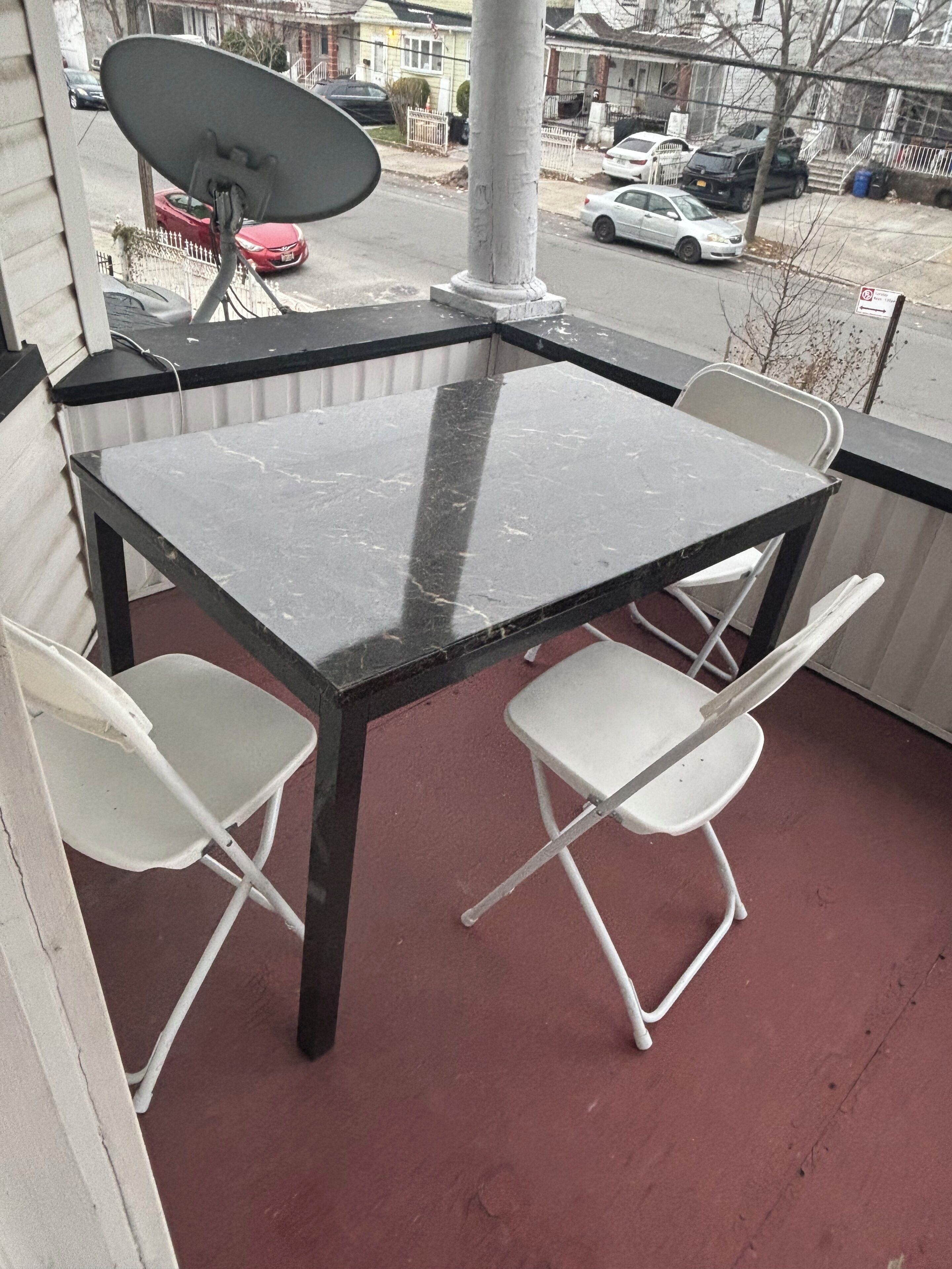 A balcony with a table and four chairs, a satellite dish, and a view of parked cars and houses.