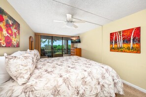 A bedroom with a large bed, a ceiling fan, a TV, and two paintings. - Kona Pacific A202 - Oceanview Kona Condo | Walk to Town & Ironman (Kailua Kona)