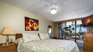 Quarto de hotel com cama, mesa de cabeceira, abajur, quadro, televisão e vista para palmeiras.