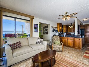 A living room with a sofa, armchair, and coffee table, a kitchen with wooden cabinets and a countertop, and a ceiling fan. - Kona Pacific A202 - Oceanview Kona Condo | Walk to Town & Ironman (Kailua Kona)