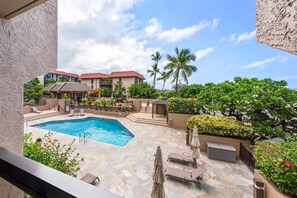 A swimming pool with lounge chairs and umbrellas, surrounded by greenery and buildings with red roofs. - Kona Pacific A202 - Oceanview Kona Condo | Walk to Town & Ironman (Kailua Kona)