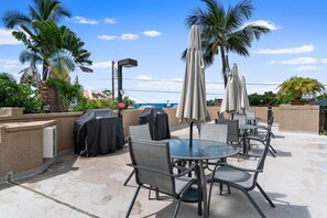 An outdoor patio with round tables and chairs, covered by umbrellas, surrounded by palm trees. - Kona Pacific A202 - Oceanview Kona Condo | Walk to Town & Ironman (Kailua Kona)