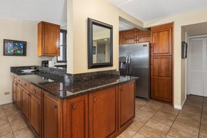 A kitchen with wooden cabinets, a granite countertop, a stainless steel refrigerator, and a large mirror. - Kona Pacific A202 - Oceanview Kona Condo | Walk to Town & Ironman (Kailua Kona)