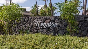 Un muro de piedra con el nombre "Kanaloa" en escritura cursiva, rodeado de vegetación exuberante y palmeras.