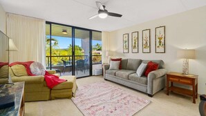 A living room with a sofa, a smaller couch, a coffee table, a patterned rug, a ceiling fan, and a balcony with a view. - Kona Pacific B411 - Garden View Kona Condo | Walk to Town & Ironman (Kailua Kona)