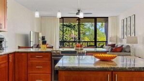 A kitchen with a large island, wooden cabinets, a dining area with a sofa, and a view of greenery outside. - Kona Pacific B411 - Garden View Kona Condo | Walk to Town & Ironman (Kailua Kona)