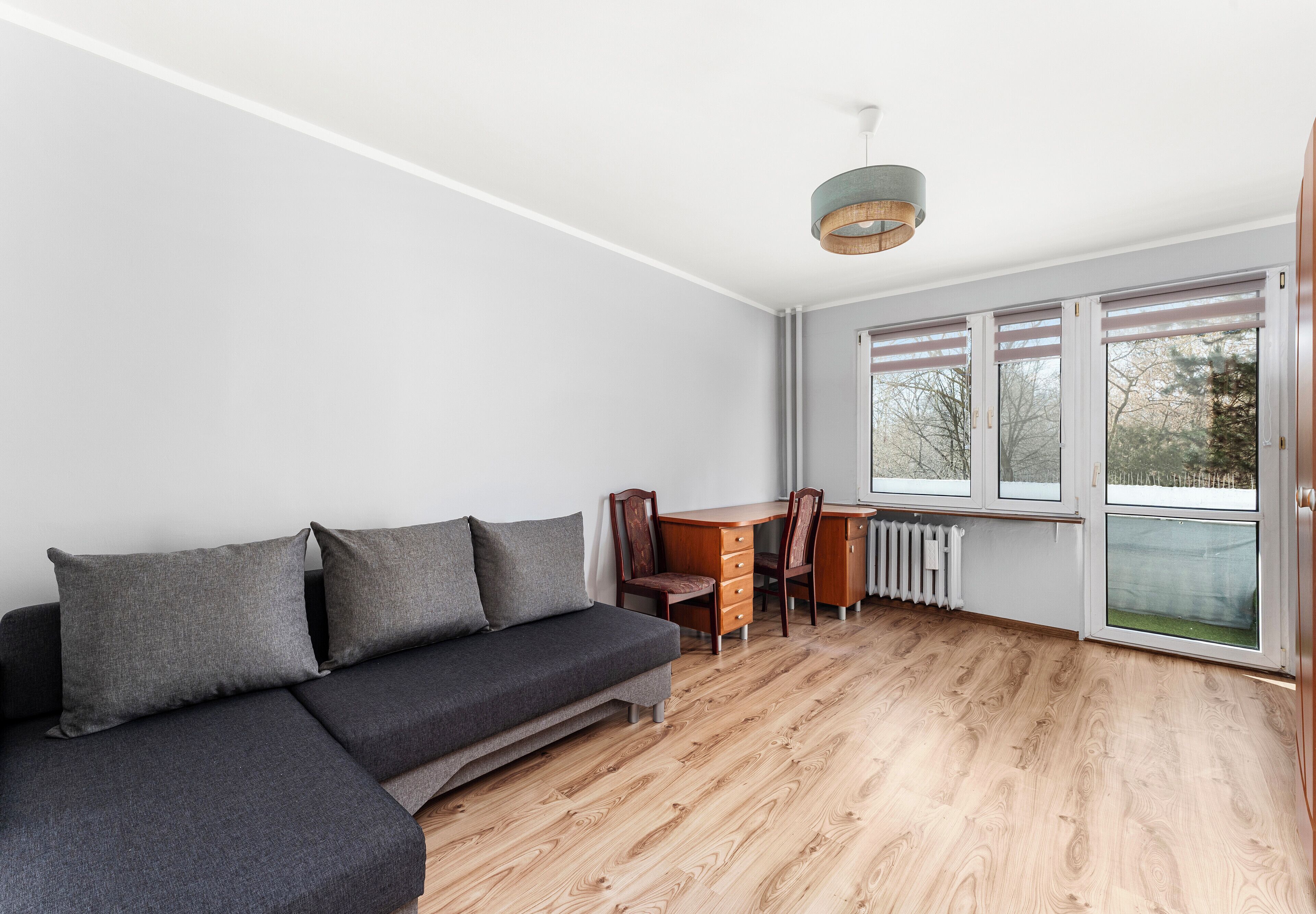 Une pièce moderne comprenant un canapé d’angle, une table à manger en bois avec des chaises, et de grandes fenêtres équipées de stores.