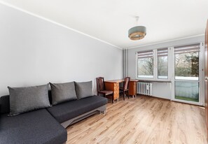 A modern room with a sectional sofa, a wooden dining table with chairs, and large windows with blinds. - Odrzańska Harmony Apartment (Kraków)