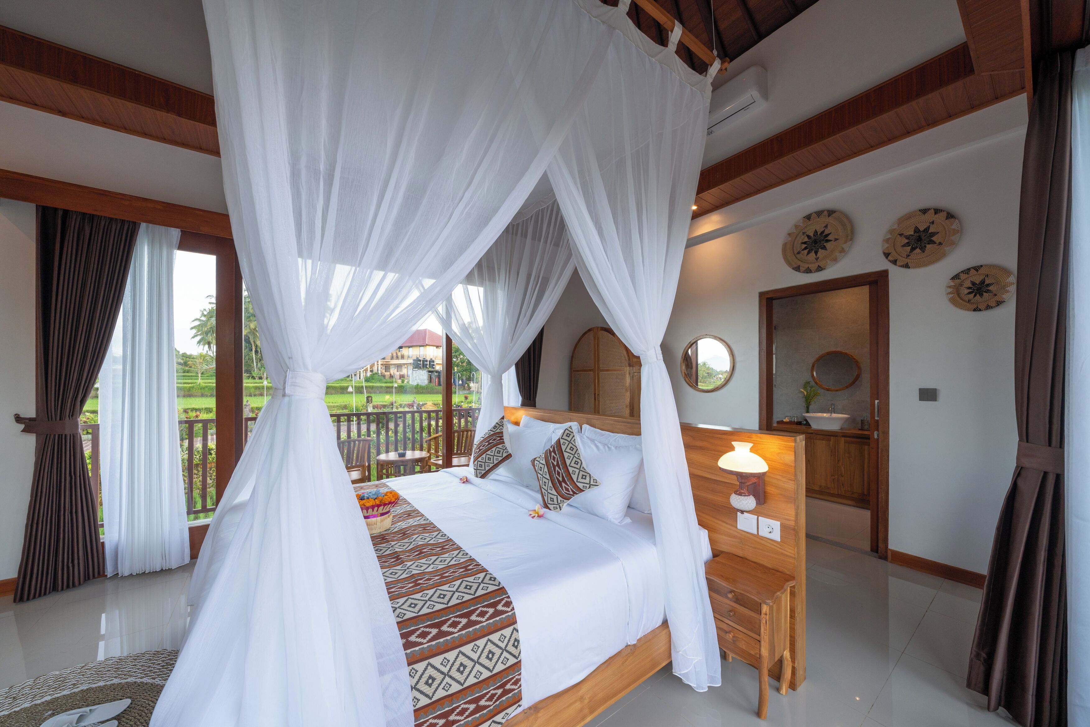 A bedroom with a canopy bed, a bedside table with a lamp, and a view of the outdoors.